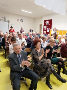 Dziadkowie i babcie uczniów klasy drugiej siedzą na krzesełkach ustawionych w sali uczniów. Dziadkowie klaszczą.