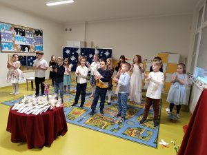 Uczniowie klasy drugiej podczas występu z okazji dnia babci i dziadka. Dzieci stoją na dywanie i klaszczą w dłonie, a przed nimi stoi okrągły stolik z upominkami dla dziadków.