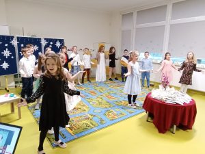 Uczniowie klasy drugiej podczas występu z okazji dnia babci i dziadka. Dzieci wykonują piosenkę w trakcie której wszyscy szeroko rozkładają dłonie. Dzieci stoją na dywanie a przed nimi stoi okrągły stolik z upominkami dla dziadków.