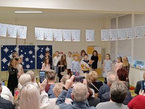 Uczniowie klasy drugiej podczas występu z okazji dnia babci i dziadka. Nad uczniami wisi girlanda z napisem "Dzień babci i dziadka". Na przeciwko siedzą na krzesełkach dziadkowie i babcie.