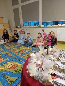 Uczniowie klasy drugiej siedzą na podłodze wzdłuż dywanu. Jedna z dziewczynek wyciąga w górę kartkę z narysowaną uśmiechniętą buźką. Po prawej stronie na zdjęciu widoczny jest fragment okrągłego stołu przykrytego czerwonym materiałem z upominkami dla dziadków.