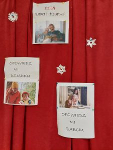 Na czerwonym materiale zawieszone są trzy białe gwiazdki oraz trzy kartki ze zdjęciami uczniów z dziadkami. Pod każdym ze zdjęć umieszczone są napisy: "DZIEŃ BABCI I DZIADKA", "OPOWIEDZ MI DZIADKU", "OPOWIEDZ MI BABCIU".