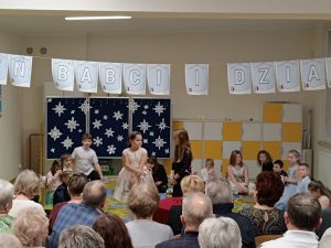 Uczniowie klasy drugiej podczas występu z okazji dnia babci i dziadka. Nad uczniami wisi girlanda z napisem "Dzień babci i dziadka". Na przeciwko siedzą na krzesełkach dziadkowie i babcie.