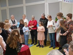 W rzędzie pod oknem stoją bacie i dziadkowie. Przed nimi stoi troje uczniów klasy drugiej. Dzieci spoglądają na trzymane w dłoniach karteczki.