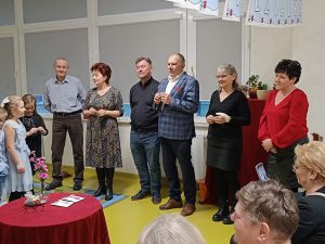 Sześcioro babć i dziadków stoi w rzędzie pod oknem. Na przeciwko nich widoczny jest okrągły stolik przykryty czerwonym materiałem.