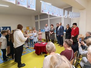 Sześcioro babć i dziadków stoi w rzędzie pod oknem. Na przeciwko nich stoi wychowawczyni z pudełkiem z upominkami oraz uczniowie klasy. Pomiędzy nimi ustawiony jest okrągły stolik przykryty czerwonym materiałem.