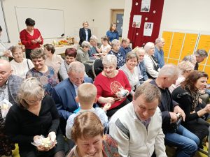 Dziadkowie i babcie siedzą na krzesełkach ustawionych w sali klasy drugiej i oglądają otrzymane upominki.