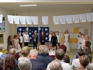 Uczniowie klasy drugiej podczas występu z okazji dnia babci i dziadka. Nad uczniami wisi girlanda z napisem "Dzień babci i dziadka". Na przeciwko siedzą na krzesełkach dziadkowie i babcie.