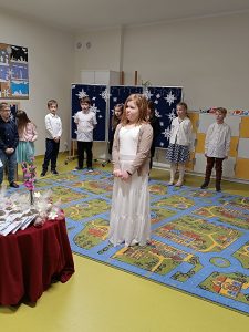 Uczniowie klasy drugiej stoją w półokręgu wzdłuż dywanu. Po środku stoi dziewczynka ubrana w białą długą sukienkę.