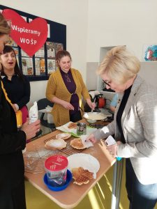Przy stoliku stoją dwie mamy i pani dyrektor. Na stole leżą gofry upieczone przez mamy. Pani dyrektor bierze do ręki jeden z nich. W tle widać przyczepione na ścianę czerwone serduszko z napisem "Wspieramy WOŚP".
