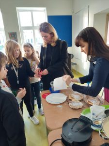 Przy stoliku z goframi stoi jedna z mam i pani nauczycielka. Mama trzyma w dłoni gofra przykrytego chusteczką, Nauczycielka daje dziewczynce stojącej obok pieniądze.