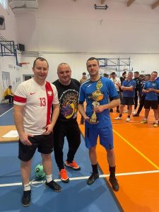 Troje zawodników biorących udział w rywalizacji pozuje do wspólnego zdjęcia. Jeden z nich trzyma puchar. W tle widać stojących innych graczy.