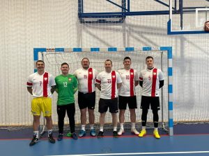 Sześcioro mężczyzn pozuje do wspólnego zdjęcia stojąc w świetle bramki. Pięcioro z nich ma jednakowe białe koszulki z czerwonym podłużnym paskiem. Jeden jest ubrany w zieloną koszulkę.