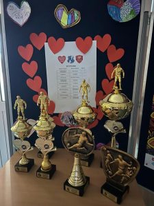 Zdjęcie ustawionych na stoliku pucharów przygotowanych dla zwycięzców spotkania. Za nimi na tablicy wywieszona jest kartka z rozgrywkami drużyn a wokół niej czerwone małe serduszka ułożone w duże serce.