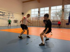 Zdjęcie wykonane w trakcie rozgrywki. Między nogami jednego z zawodników przelatuje piłka. Przed nim stoi dwóch zawodników z przeciwnej drużyny.