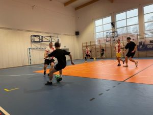 Zdjęcie wykonane podczas jednej z rozgrywek. Jeden z zawodników kopie zieloną piłkę a mężczyzna biegnący z naprzeciwka próbuje ją zatrzymać.