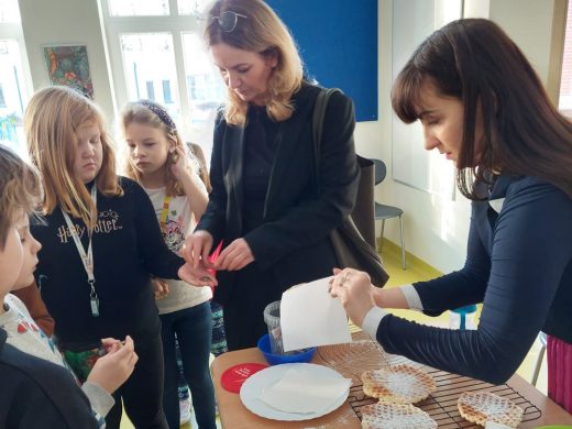 Przy stoliku z goframi stoi jedna z mam i pani nauczycielka. Mama trzyma w dłoni gofra przykrytego chusteczką, Nauczycielka daje dziewczynce stojącej obok pieniądze.
