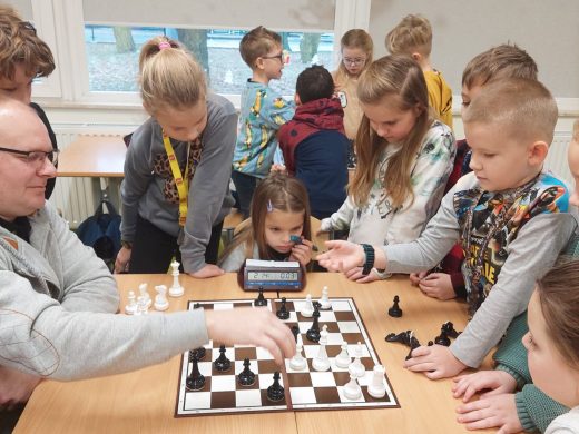 Uczniowie klasy drugiej stoją wokół stołu z rozstawioną szachownicą. Przy stole siedzi pan Kamiński przestawiający pionki na szachownicy.