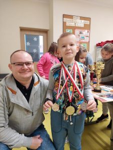 Uczeń klasy drugiej stoi z zawieszonymi na szyi medalami. Obok niego kuca pan Kamiński. W tle widać innych uczniów przyglądających się nagrodom bohatera spotkania.
