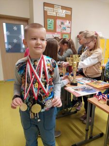 Uczeń klasy drugiej stoi z zawieszonymi na szyi medalami. W tle widać innych uczniów przyglądających się nagrodom bohatera spotkania.