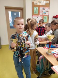 Uczeń klasy drugiej stoi trzymając w dłoniach dwa puchary. W tle widać innych uczniów przyglądających się nagrodom bohatera spotkania.