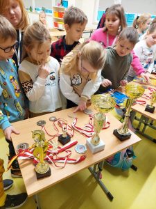 Uczniowie klasy drugiej oglądają ułożone na stoliku medale i puchary.