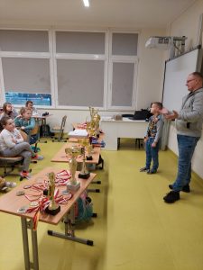 Pan Kamiński wraz z jednym z uczniów stoi przed stołami, na których rozłożone są jego puchary i medale zdobyte w turniejach szachowych.