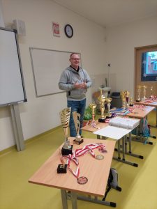 Pan Kamiński stoi przed stołami, na których rozłożone są jego puchary i medale zdobyte w turniejach szachowych.
