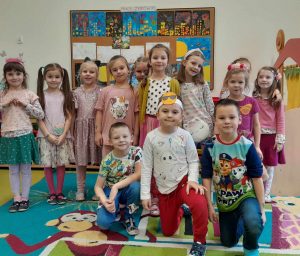 Uczniowie oddziału przedszkolnego pozują do wspólnego zdjęcia. Dziewczynki stoją z tyłu a przed nimi na kolanach kucają chłopcy. Jeden z nich ma na głowie opaskę z uśmiechniętą buźką w okularach przeciwsłonecznych.