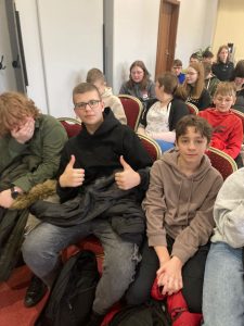 Uczniowie klasy ósmej siedzą na czerwonych krzesłach w rzędach za sobą. W pierwszym rzędzie siedzi troje uczniów - jeden z nich podnosi w górę oba kciuki.