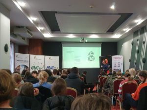 Pan Robert Baca podczas wystąpienia motywującego przygotowanego dla uczestników projektu matma^2. Za nim na rzutniku wyświetlona jest grafika przedstawiająca niezadowolonego ucznia. Obok stoją banery prezentujące projekty Matma^2 i Matura+ oraz wydawnictwo Elitmat.