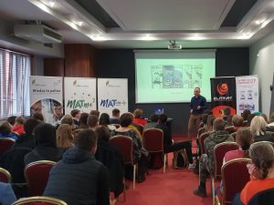 Pan Robert Baca podczas wystąpienia motywującego przygotowanego dla uczestników projektu matma^2. Za nim na rzutniku wyświetlony jest komiks. Obok stoją banery prezentujące projekty Matma^2 i Matura+ oraz wydawnictwo Elitmat.