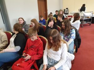 Uczniowie klasy ósmej siedzą w czteroosobowych rzędach na czerwonych fotelach. Część z nich ma przed sobą telefony.