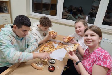 Czworo uczniów klasy szóstej siedzi przy wspólnym stole i je pizze. Chłopcy mają spuszczone w dół głowy a dziewczynki się uśmiechają patrząc wprost w obiektyw i prezentując trzymane w dłoniach pizze.
