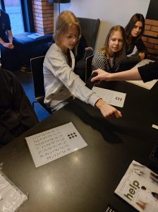 Trzy uczennice siedzą przy długim ciemnym stole. Jedna z dziewczynek wyciąga przed siebie ręką, do której ma przykładane czarne urządzenia. Uczennica i jej koleżanki patrzą na to urządzenie.