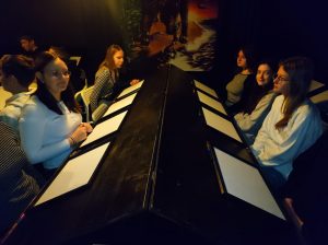 Uczennice klasy siódmej siedzą w dwóch rzędach na przeciwko siebie. Między nimi stoi stół z pochylonymi blatami. Przed każdą z dziewczyn leży biała kartka papieru.