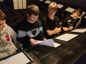 Czterech uczniów klasy siódmej siedzi przy stole z pochyłym blatem. Przed nimi leżą białe kartki papieru. Jeden z uczniów podnosi swoją kartkę.