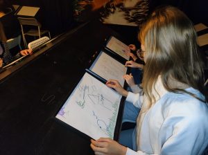 Trzy uczennice klasy siódmej siedzą przy stole z pochyłym blatem. Przed dziewczynkami leżą kartki papieru z rysunkami przez nie wykonanymi.