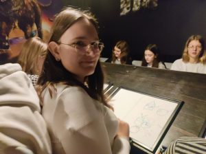 Uczennice klasy siódmej siedzą w dwóch rzędach przed sobą. Między nimi ustawiony jest stół z pochylonymi blatami. Jedna z uczennic uśmiecha się w stronę aparatu. Przed nią leży kartka papieru z wykonanym przez nią rysunkiem.