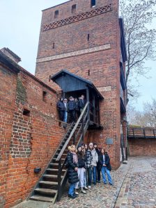 Uczniowie klasy siódmej pozują do wspólnego zdjęcia przy krzywej wieży w Toruniu. Dziewczynki stoją na dole a chłopcy na piętrze przed wejściem do wieży.