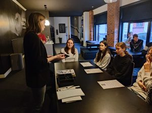 Uczennice klasy siódmej siedzą przy stole, na którym leżą kartki papieru. Przed nimi stoi pracownica muzeum.