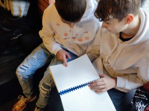 Dwóch uczniów klasy siódmej przygląda się notesowi z jasnymi kartkami, który trzyma jeden z nich.