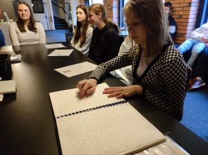 Uczennice klasy piątej siedzą przy długim stole. Dziewczynka widoczna na pierwszym planie przygląda się leżącemu przed nią skoroszytowi zapisanemu w języku braille'a. Dziewczynka przesuwa palcem po napisach.