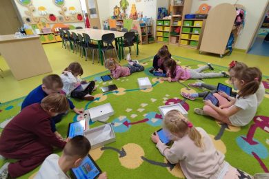 Uczniowie oddziału przedszkolnego leżą lub siedzą na dywanie w okręgu. Dzieci trzymają w dłoniach tablety i patrzą w ich ekrany.