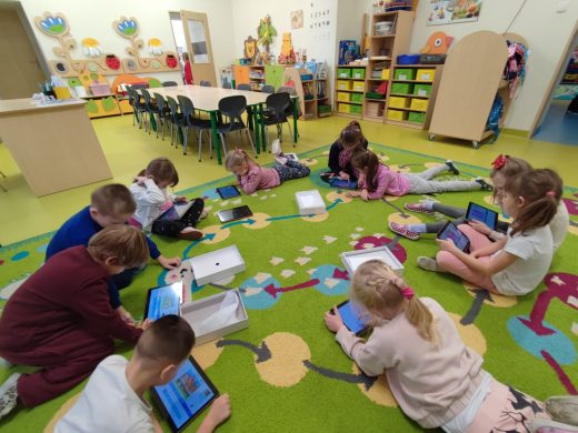 Uczniowie oddziału przedszkolnego leżą lub siedzą na dywanie w okręgu. Dzieci trzymają w dłoniach tablety i patrzą w ich ekrany.