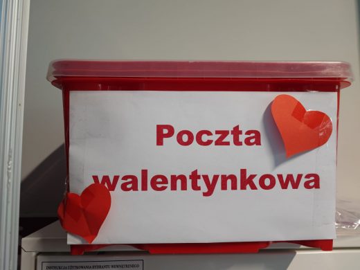 Zdjęcie czerwonego pudełka z białą kartką z nadrukowanym czerwonym napisem :"Poczta Walentynkowa". Pudełko ozdobiono dwoma czerwonymi serduszkami przyklejonymi do kartki.