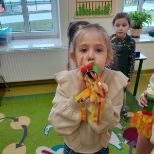 Na zdjęciu widoczna jest uczennica oddziału przedszkolnego, która trzyma przy ustaw wykonaną przez siebie dmuchawkę przypominającą smoka wawelskiego z długim ogonem wyciętym z krepy. W tle widać stojącą inną uczennicę.