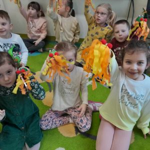 Uczniowie oddziału przedszkolnego siedzą na dywanie. Dzieci trzymają w dłoniach wykonane przez siebie dmuchawki przypominające smoka wawelskiego.