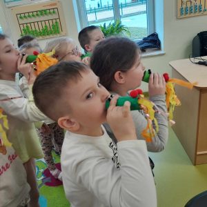 Uczniowie oddziału przedszkolnego stoją w grupie na dywanie. Przy ustach trzymają dmuchawki przypominające smoka wawelskiego z długimi ogonami.