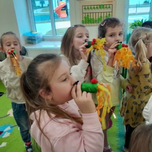 Cztery dziewczynki z oddziału przedszkolnego stoją na dywanie. Przy ustach trzymają dmuchawki przypominające smoka wawelskiego z długimi ogonami.